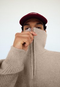 Maglione beige in maglia con collo alto e mezza cerniera, caratterizzato da una trama a coste. Cappellino rosso con logo bianco indossato sopra.