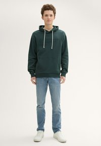 Ciemnozielona bluza z kapturem, z sznurkiem, kieszenią z przodu i wytłoczonym logo. Skierowana z jasnoniebieskimi dżinsami i białymi sneakersami. Gładka tekstura.