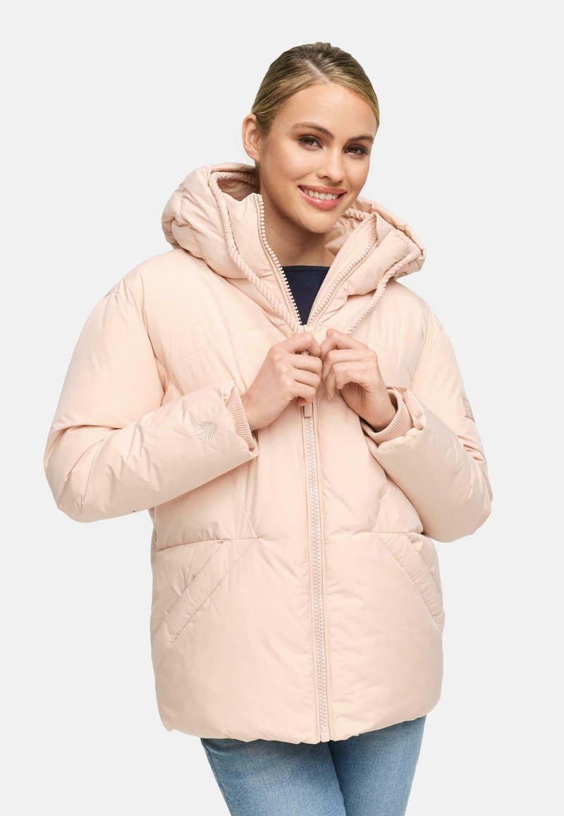 Vrouw met een glimlach, gekleed in een lichtroze gewatteerde jas met een capuchon, die half dicht is geritst, gecombineerd met blauwe jeans tegen een effen achtergrond.