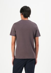 Mann mit kurzem dunklem Haar, der ein einfarbiges braunes T-Shirt mit kurzen Ärmeln und dunkle Hosen trägt, steht mit dem Rücken zu einem weißen Hintergrund.
