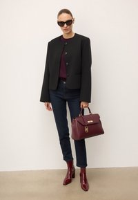 Črna skrajšana jakna s gumbi, bordo top, oprijete temno jeans hlače in sijoči bordo škornji s peto. Drži strukturirano bordo torbico.