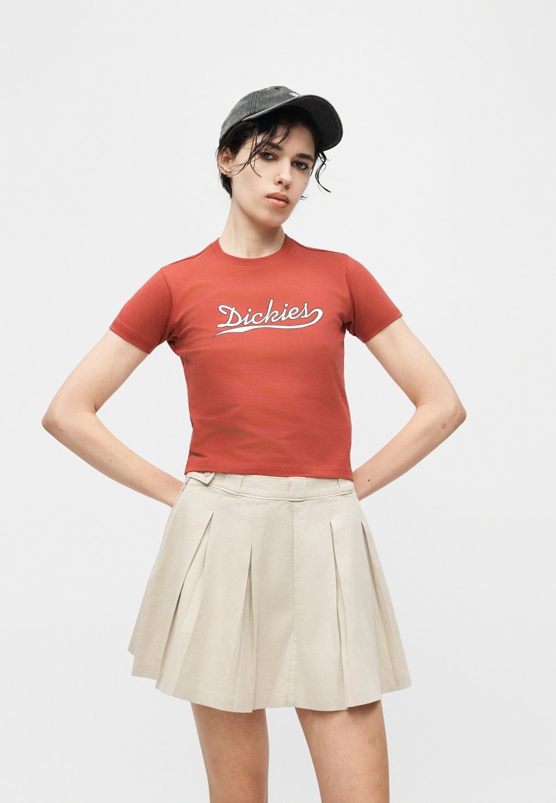 Giovane donna con maglietta rossa Dickies, gonna beige plissettata e cappellino nero, in piedi con le mani sui fianchi davanti a uno sfondo bianco.
