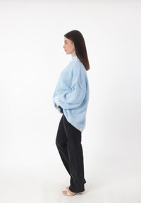 Lichtblauwe oversized gebreide trui met een geribbelde halslijn, samen met zwarte rechte jeans, gezien vanuit de zijkant.