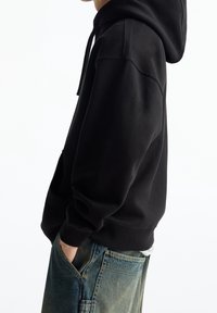 Svart luvtröja med känguruficka, avslappnad passform och ribbade muddar. Bärs med ljusblå baggy denimbyxor.