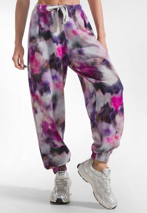 Persona che indossa pantaloni della tuta tie-dye loose fit in rosa, viola, nero e grigio con coulisse bianca e sneakers argentate.