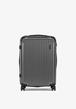 Szara walizka z twardą skorupą, z pionowymi żebrowaniami, czterema kółkami i górnym uchwytem, stojąca pionowo na białym tle.