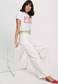 Biała bawełniana koszulka z kolorową grafiką, białe spodnie z wysokim stanem i jasnoszary pasek. Czerwone baletki dopełniają stylizację.