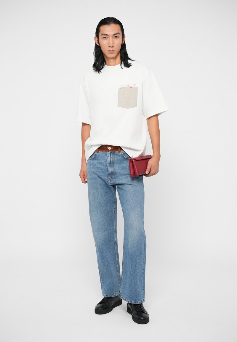 Mann mit langen schwarzen Haaren, der ein weißes T-Shirt mit beigefarbenem Taschen, blaue Jeans und schwarze Schuhe trägt und eine rote Clutch vor einem weißen Hintergrund hält.