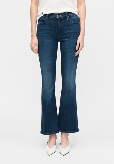 Γυναίκα που φοράει σκούρα μπλε τζιν καμπάνα, άσπρα ψηλοτάκουνα παπούτσια με μυτερή μύτη και άσπρο τοπ, στέκεται μπροστά σε απλό λευκό φόντο.