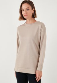 Maglietta beige a maniche lunghe con scollatura rotonda e spalle scese, caratterizzata da una texture liscia e una vestibilità morbida, abbinata a jeans scuri.