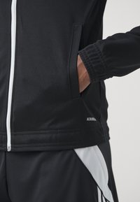 Svart sportjacka med dragkedja, sidofickor och ribbade muddar, med vita detaljer och ett texturerat tyg.