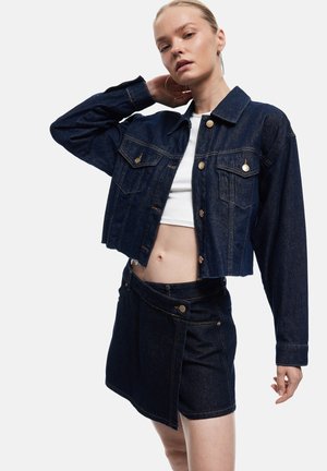 Frau trägt dunkelblaue, kurz geschnittene Jeansjacke und passenden Rock mit asymmetrischer Vorderseite, posiert mit einer Hand hinter dem Kopf.