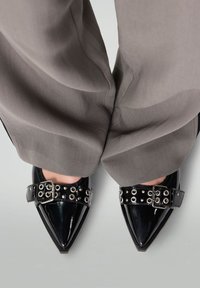 Zwarte gepatineerde leren schoenen met puntige neuzen en een glanzende afwerking, voorzien van een zwarte band versierd met zilveren oogjes en een metalen gesp.