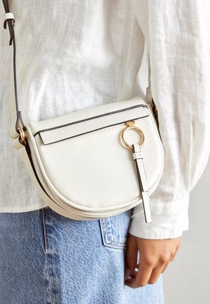 Biała skórzana torebka crossbody ze złotym pierścieniem, noszona na białej lnianej koszuli i niebieskich dżinsach.