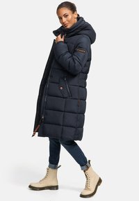 Manteau long matelassé bleu marine avec capuche, doté d'une fermeture éclair et de boutons-pression, de deux poches latérales, et d'un tissu texturé avec un motif matelassé.