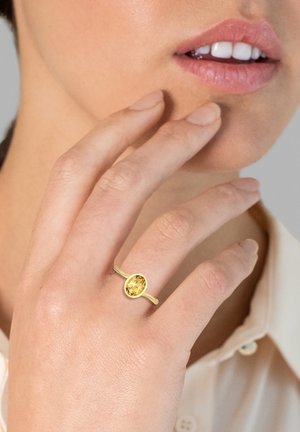 Goldring mit einem ovalen gelben Edelstein, auf einem dünnen Band gefasst, an einer Hand getragen, mit einer blass gefärbten Bluse im Hintergrund.