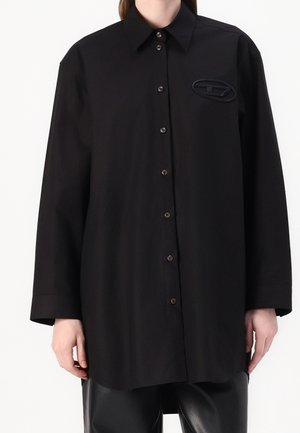 Personne portant une chemise noire à manches longues avec des boutons, un col pointu et un écusson ovale sur le côté gauche de la poitrine.