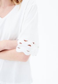 Top in cotone bianco con scollatura a V e maniche corte, caratterizzato da ritagli ondulati ai polsini. Texture liscia, vestibilità casual.