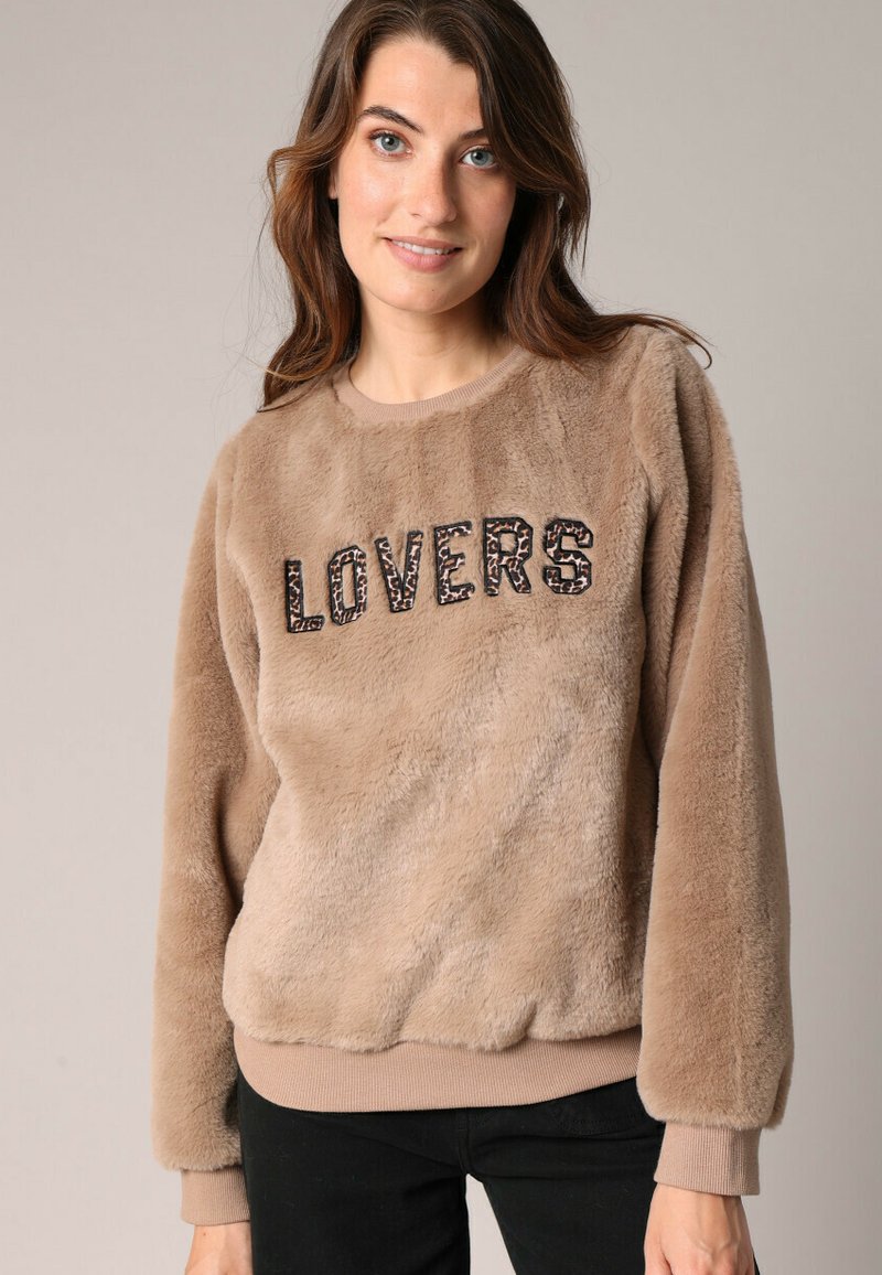 Sweatshirt en fausse fourrure beige avec le texte "LOVERS" en impression foncée, col rond, ourlet et poignets côtelés, et texture pelucheuse. Design décontracté avec des manches longues.