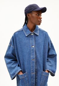 Oversized blue denim jacket with a collar, front buttons, and large pockets. Accompanied by a navy cap. Textured fabric and relaxed fit.