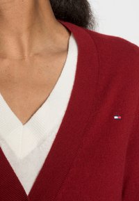 Červený kardigan s výstřihem do V, přes bílý svetr s výstřihem do V. Obsahuje jemný detail značky ve formě malé logové visačky. Měkká textura.