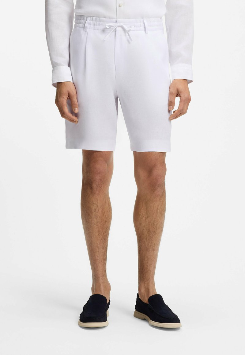 Homme portant un short blanc à cordon, une chemise blanche à manches longues et des chaussures noires sans lacets, debout avec les bras détendus le long du corps.