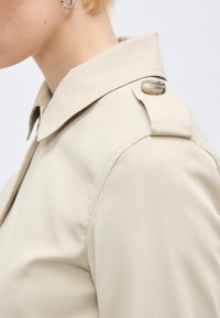 Spalla di trench beige con dettaglio bottone sull'epaulet su una persona con capelli corti biondi e orecchino a cerchio d'argento.
