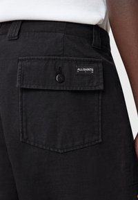 Black fabric shorts feature a back pocket with a flap, button closure, and a label. The texture is smooth, and the design is simple.