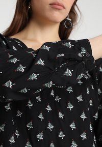 Blouse en tissu noir avec motifs répétitifs de sapins de Noël verts avec accents rouges. Manches longues froncées avec détails boutonnés.