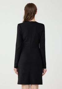 Czarna sukienka z długim rękawem, dopasowany krój, sięgająca do kolan z przeszyciami z tyłu. Gładka faktura i jednolity kolor, bez wzorów i ozdób.