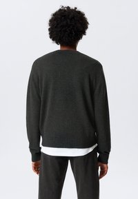 Mørkegrønn genser, strikket tekstur, rund halslinning, ribbestrikkede mansjetter og nederkant, parret med svarte jeans, synlig etikett nederst på ryggen.