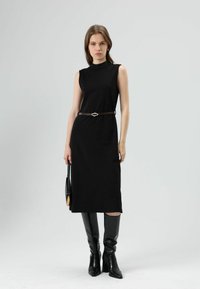 Zwarte mouwloze midi-jurk met hoge halslijn, taille aangesnoerd door een bruine riem met een decoratieve gesp; gecombineerd met zwarte kniehoge laarzen.