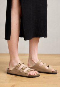Sandálias de três tiras na cor tan com fechos dourados, sola de cortiça e parte superior em couro liso, usadas com um vestido preto canelado.