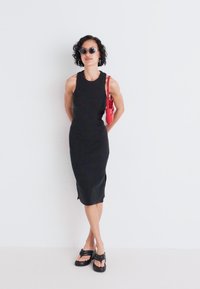 Zwarte mouwloze midi-jurk met een slim fit. Gladde textuur en zijsplitten. Gecombineerd met zwarte sandalen en een rode handtas.