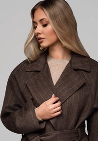 Abrigo de lana marrón a rayas con un amplio cuello y cinturón, llevado sobre un suéter ligero. Lleva pendientes de oro. El cabello está peinado en ondas sueltas.