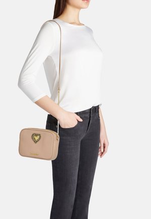 Borsa a tracolla sintetica beige con un motivo a forma di cuore e tracolla a catena, dotata di chiusura con zip. Indossata con una camicia bianca e jeans neri.