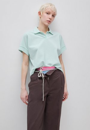 Osoba z krótkimi blond włosami, ubrana w jasnoniebieską koszulkę polo włożoną do ciemnobrązowych spodni z widocznym różowym pasem i białym sznurkiem.