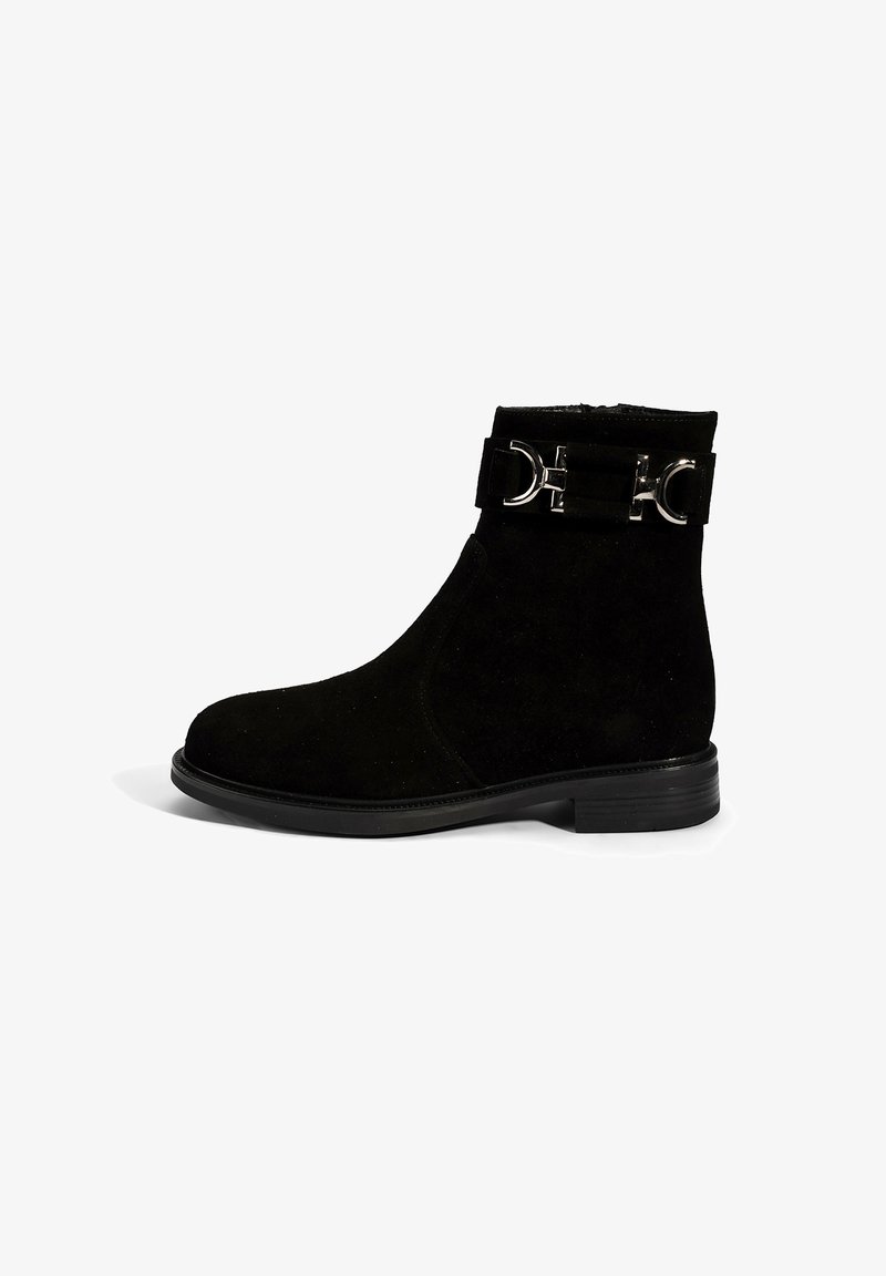 Bottines en suède noir avec un bout arrondi, un faible talon, et une sangle décorative avec des accents en métal argenté. Texture lisse et design ajusté.