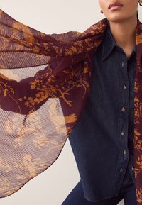Écharpe en mousseline bordeaux avec motifs floraux et oiseaux orange, drapée sur une chemise en jean. Tissu léger avec une finition texturée.