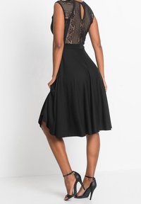Černé šaty s krajkovým živůtkem, bez rukávů, s rozšířenou sukní a kapsami. Doplněné černými vysokými sandály. Detail ve tvaru klíčové díry na zádech.