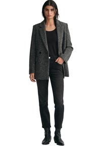 Grå herringbone-blazer med spetsiga slag, en knapp och sidofickor, bärs över en svart topp och matchas med svarta jeans och ankelboots.