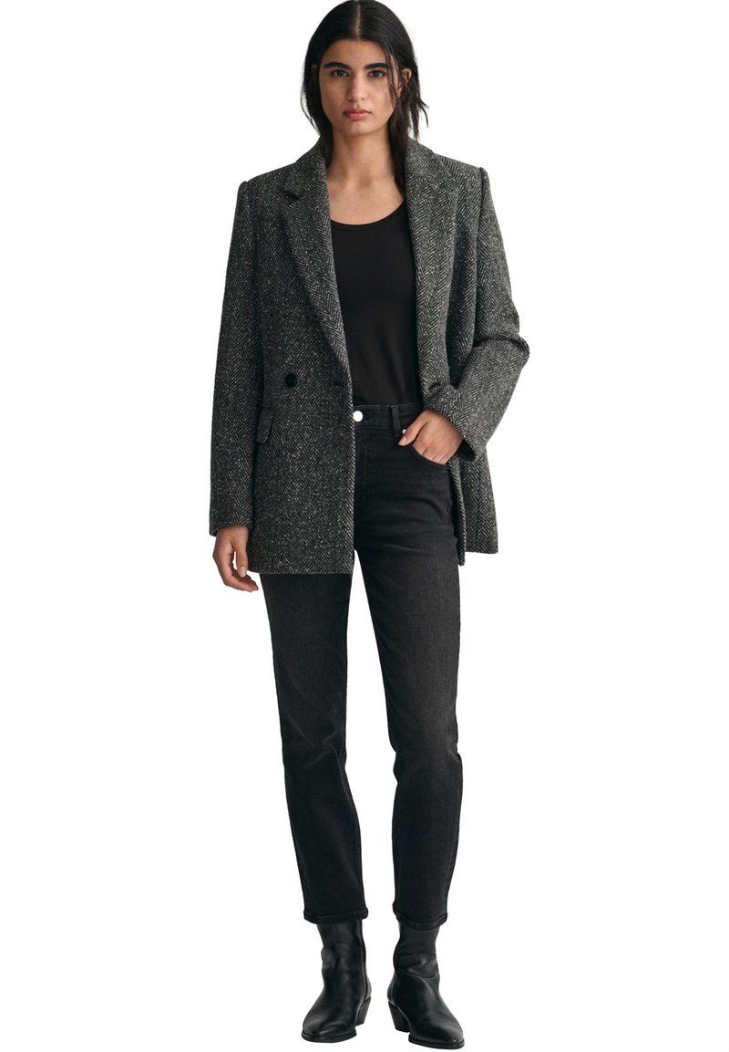 Grå herringbone-blazer med spetsiga slag, en knapp och sidofickor, bärs över en svart topp och matchas med svarta jeans och ankelboots.