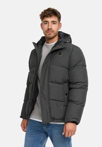 Man in een donkergrijze pufferjas met capuchon, lichtgrijze sweater en blauwe jeans, staand tegen een witte achtergrond.