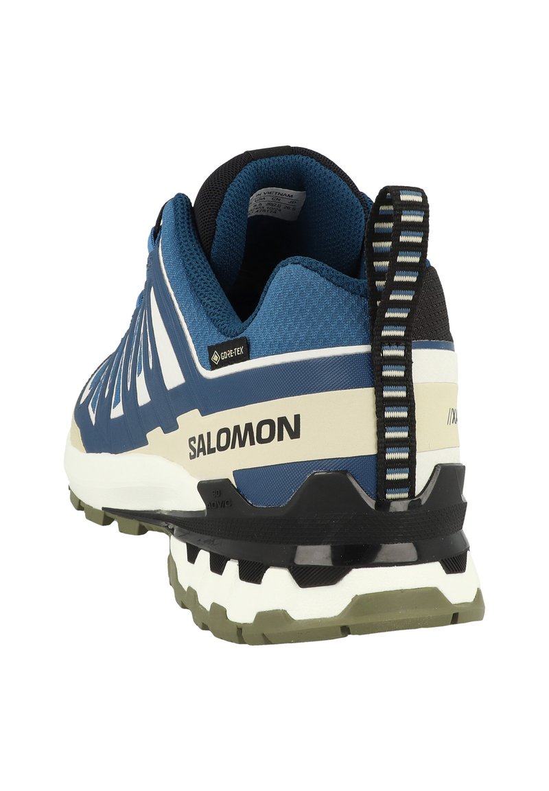 Chaussure de sport bleue et noire avec une tige texturée, des accents blancs, une semelle en caoutchouc avec des crampons multidirectionnels, et une languette au talon.