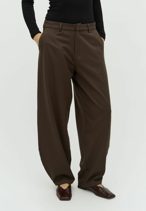 Kobieta ubrana w luźne, ciemnobrązowe spodnie, czarną bluzkę z długim rękawem oraz ciemnobrązowe, szpiczaste baleriny, stojąca z rękami w kieszeniach.