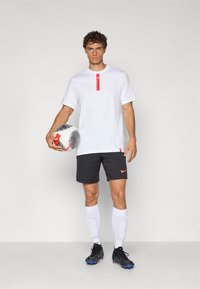 Hvid t-shirt med rød label, sorte shorts med orange Nike-logo, hvide sokker, sorte og blå fodboldstøvler, holder en sort og hvid fodbold.