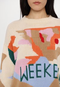 Femme portant un pull en tricot avec des formes abstraites en orange, rose, beige et bleu, montrant partiellement le mot "WEEKEND" en lettres vertes.