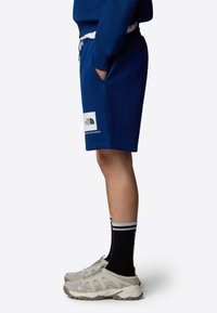 Pantalones cortos de algodón azul marino con cintura elástica y bolsillos laterales; presenta un parche de logo blanco en la pierna. Llevado con zapatos grises y calcetines negros.