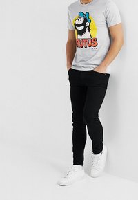 Szary t-shirt z kolorowym, kreskówkowym wzorem przedstawiającym postać z brodą, zestawiony z czarnymi, dopasowanymi spodniami oraz białymi trampkami.