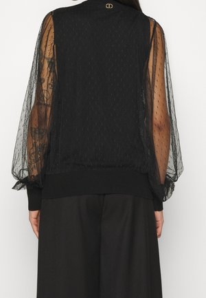 Mujer con blusa negra transparente de manga larga con patrón de puntos y pantalones negros, vista desde atrás sobre un fondo blanco.